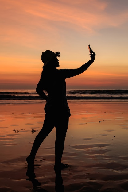 Silueta de mujer tailandesa con smartphone en la playa en el atardecer