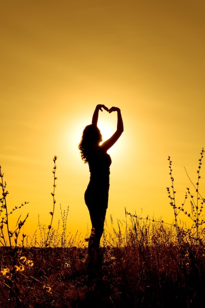 Silueta de una niña al atardecer en un campo sosteniendo sus manos sobre su cabeza en forma de corazón