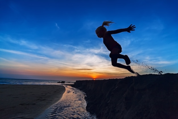 Silueta de la niña saltando alto en el aire desde un acantilado de arena en la playa al atardecer, cielo colorido