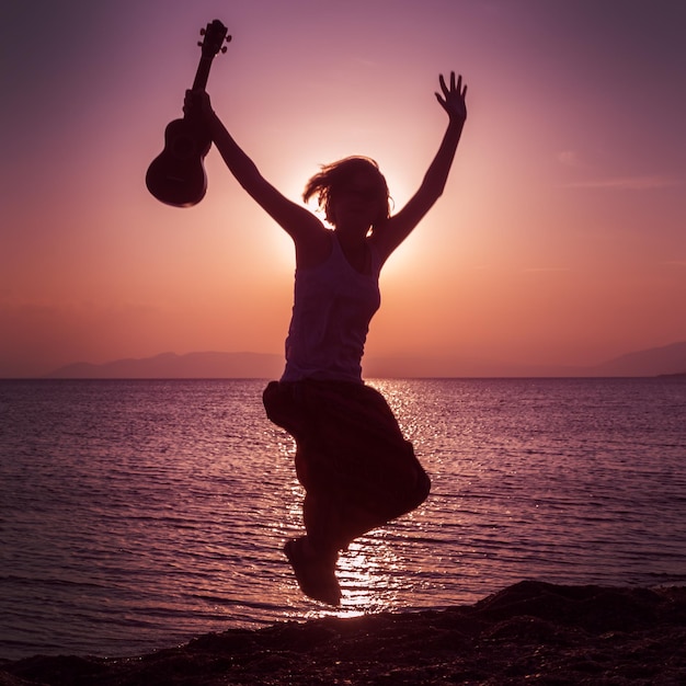 Silueta de niña saltando con las manos arriba con ukelele en la playa al atardecer