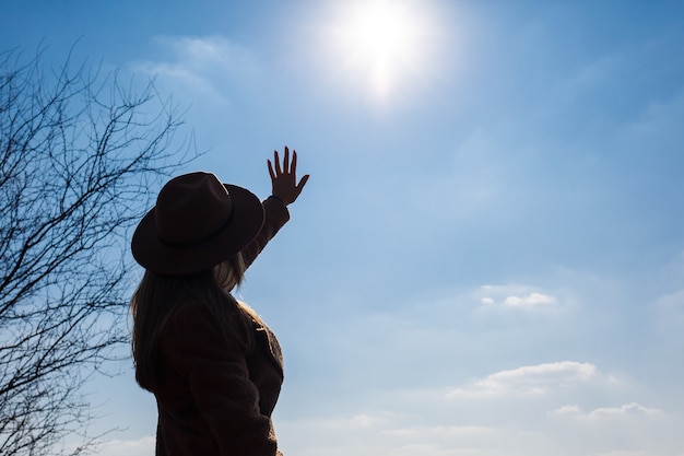 Silueta de una niña con sombrero y abrigo sobre un fondo de cielo con nubes y el sol.