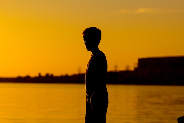 Foto silueta de un niño parado cerca del río al atardecer.