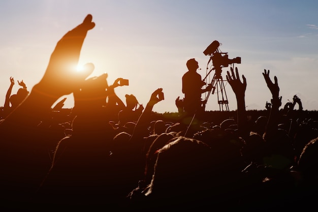 Silueta de operador de cámara filmando un concierto de rock en vivo de verano, fanáticos alrededor de las manos levantadas