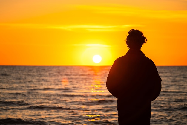 Silueta oscura de un hombre de pie junto al mar al atardecer dorado. Adolescente solitario mirando un sol poniente.