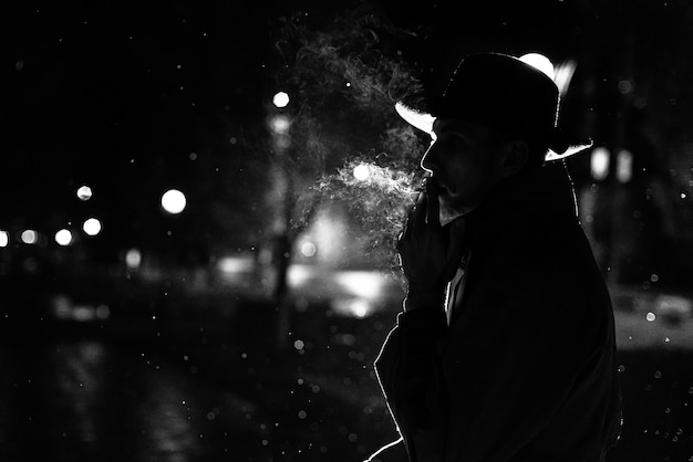 Silueta oscura de un hombre con sombrero fumando un cigarrillo bajo la lluvia en una calle nocturna al estilo de Noir