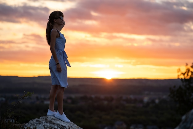 Foto silueta oscura de una mujer joven de pie sobre una piedra disfrutando de la vista del atardecer al aire libre en verano.