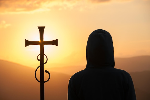 Foto silueta oscura de una persona con capucha y cruz cristiana contra el sol en el fondo