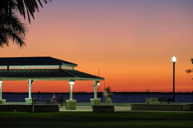 Silueta oscura de personas descansando bajo el techo de la alcoba en la orilla del mar en un parque público al atardecer