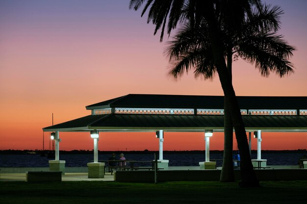 Silueta oscura de personas descansando bajo el techo de la alcoba en la orilla del mar en un parque público al atardecer