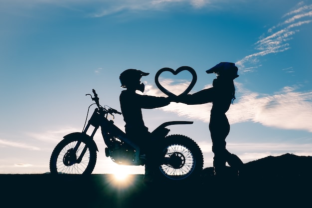 Silueta de pareja de pie y en forma de corazón en handwith una motocicleta en la puesta de sol