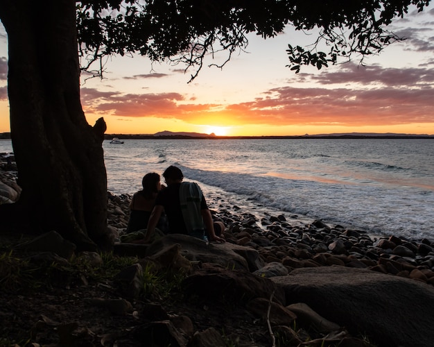 Silueta de una pareja sentada bajo un árbol mirando la puesta de sol en Noosa, Queensland, Australia