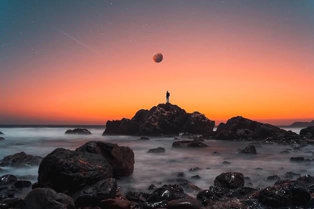 Silueta de persona en la cima de la roca con fondo de luna y estrellas sobre el océano