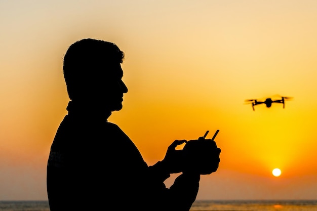 Silueta de un piloto de drones operando un drone de pie junto al mar al atardecer Vista de la silueta de un piloto de drones