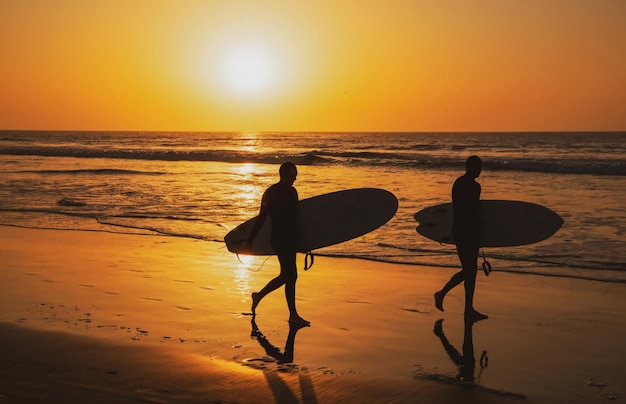 Silueta de surfistas que llevan su tabla de surf en la playa del mar al atardecer