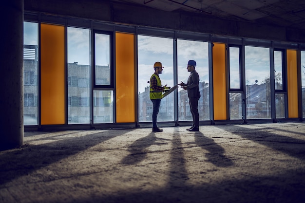 Silueta de trabajador de la construcción y arquitecto de pie junto a la ventana en el futuro centro de negocios y hablando sobre la realización del proyecto.