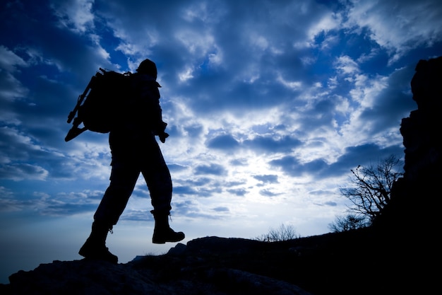 La silueta de la vista inferior de un viajero masculino con una mochila sube las montañas contra un cielo azul en nubes blancas. Concepto de desconocido y amantes del turismo de montaña