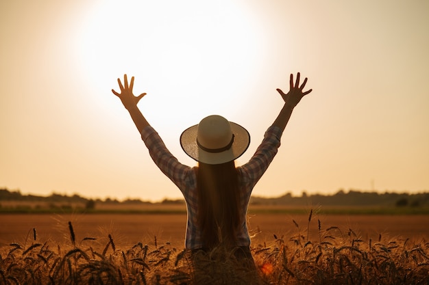 Silueta de vista trasera de mujer con sombrero en la cabeza de los agricultores con las manos levantadas al atardecer