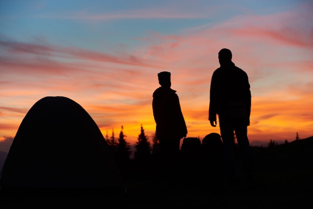 Siluetas de un hombre y una mujer de pie cerca de su tienda de campaña en la cima de una montaña impresionante puesta de sol