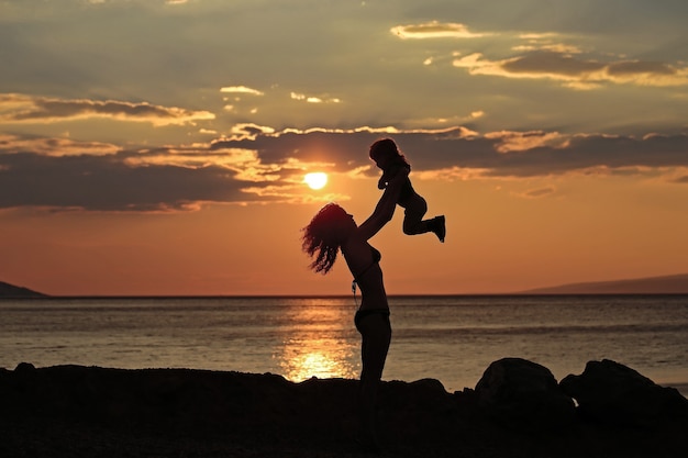 Siluetas de madre y bebé juega en la playa cerca del agua del mar o del océano en la noche o el crepúsculo día de verano al atardecer en natural