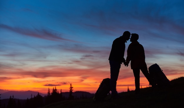 Siluetas de una pareja amorosa de excursionistas besándose en la cima de la montaña tomados de la mano