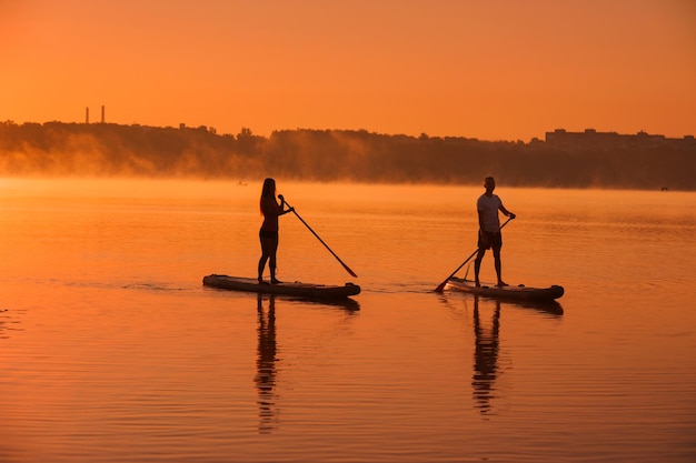 Siluetas de una pareja en tablas de SUP en el agua del lago contra el fondo del cielo rojo con recreación activa de niebla matutina