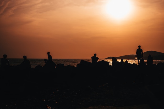 siluetas de personas descansando en la playa junto al mar contra la puesta de sol naranja