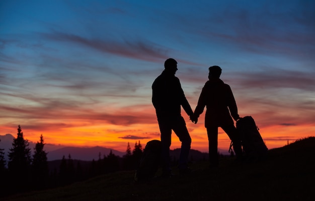 Siluetas de senderismo pareja disfrutando impresionante puesta de sol en la cima de la montaña cogidos de la mano