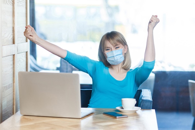 sim. Retrato de uma jovem feliz com máscara médica cirúrgica em t-shirt azul está sentado e trabalhando no laptop e comemorando sua vitória no trabalho. Conceito de trabalho, medicina e saúde indoor.