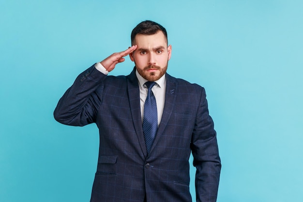 Sim senhor! Homem de negócios sério com barba vestindo terno estilo oficial segurando a mão perto da cabeça, saudando, honra e glória, militarização. Tiro de estúdio interior isolado sobre fundo azul.