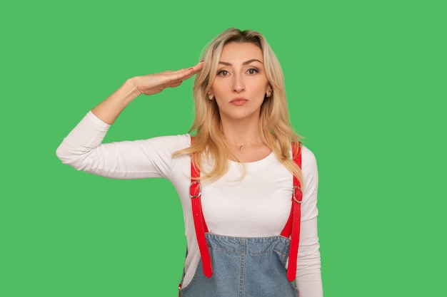 Sim senhor Retrato de mulher adulta disciplinada responsável em macacão jeans elegante saudando e ouvindo a ordem com expressão séria e atenta tiro de estúdio interior isolado em fundo verde