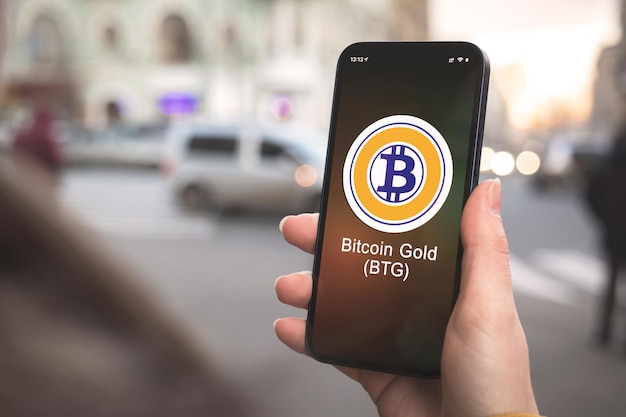 Símbolo da criptomoeda bitcoin gold btg, logotipo. conceito de negócios e financeiro. mão com smartphone, tela com close-up do ícone de criptografia