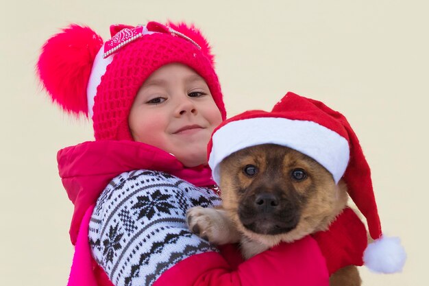 Un símbolo de la Navidad y el nuevo 2018. Feliz niña y perro en Navidad. Perro con gorra roja de Santa. Niña feliz con un pequeño cachorro en ropa de Navidad al aire libre en invierno.