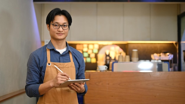 Simpático barista o camarero masculino asiático milenario sosteniendo una tableta de pie en el hermoso café