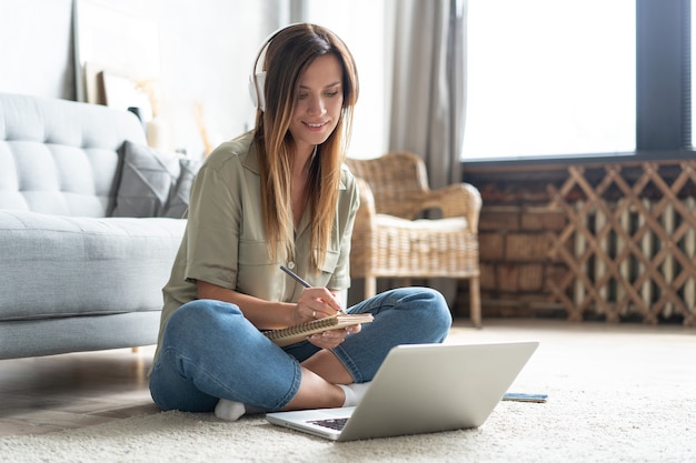 Simplemente inspirado. Mujer sonriente hermosa joven confiada que trabaja en la computadora portátil mientras que se sienta en el piso en casa.