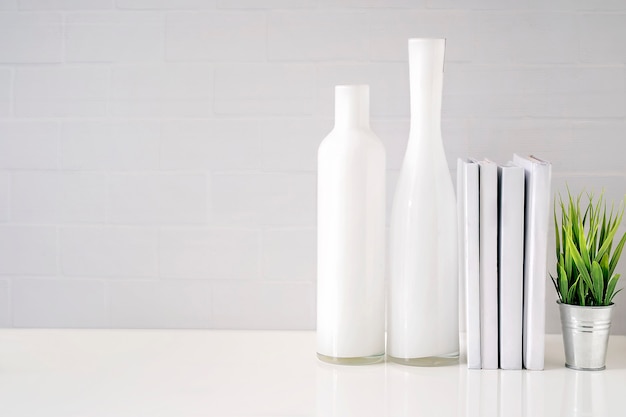 Simulacros de botellas de vidrio, libros y plantas de interior en mesa blanca con pared de ladrillo blanco