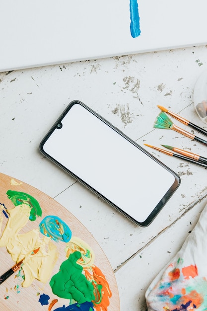 Foto simulacros de imagen de un teléfono móvil sobre una mesa blanca con una paleta de pintura y pinceles para pintar. pantalla móvil negra con espacio de copia. imagen publicitaria