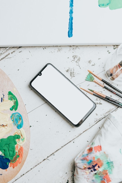 Simulacros de imagen de un teléfono móvil sobre una mesa blanca con una paleta de pintura y pinceles para pintar. Pantalla móvil negra con espacio de copia. Imagen publicitaria