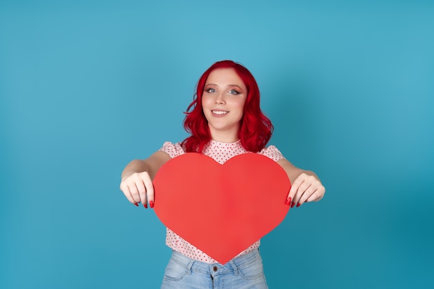 Simulacros de mujer joven feliz alegre con cabello rojo sosteniendo en dos manos un gran corazón de papel rojo