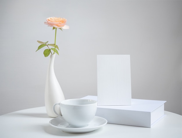 Simulacros de taza de café de tarjeta de invitación blanca y hermosas flores rosas naranjas en un moderno jarrón en mesa de madera blanca