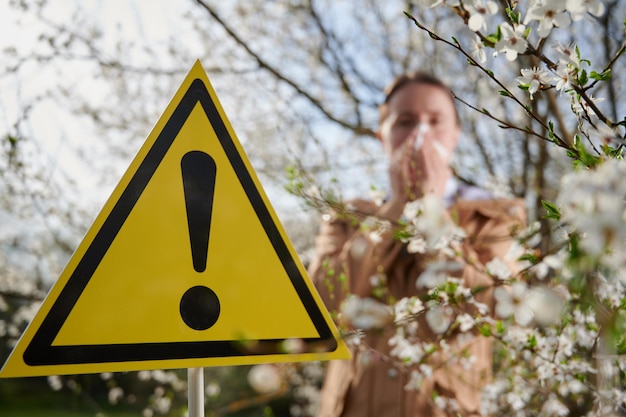 Foto sinal de atenção mulher alérgica sofrendo de alergia sazonal na primavera posando em jardim florido na primavera em fundo desfocado jovem mulher assoando o nariz conceito de alergia à primavera