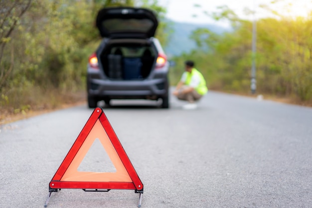 Sinal de triângulo de avaria na estrada com homem asiático preocupado reparar e trocar pneu enquanto espera o centro de serviço de seguro ou carro