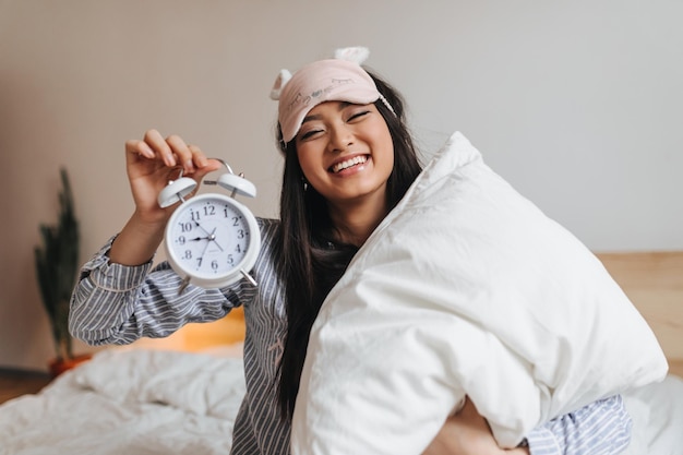 Foto sincera chica de cabello oscuro con antifaz para dormir mantiene la almohada del despertador y se ríe