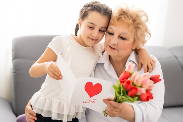 Sincera y linda nieta fuerte abrazando y besando en la mejilla su abuela de los años 60 le dio bonitas flores de primavera felicitaciones con cumpleaños, Día Internacional de la Mujer, imagen conceptual de cierre.