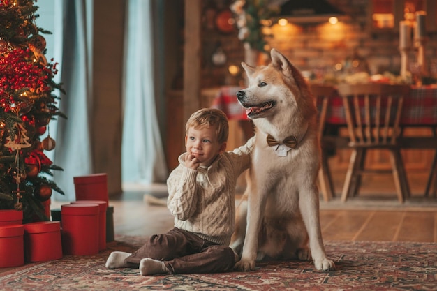 Sincero, auténtico niño feliz con suéter beige de punto abraza al perro con pajarita en casa en Navidad