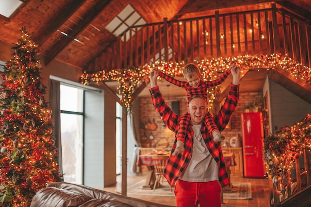 Sincero, auténtico padre feliz e hijo en pijama de cuadros rojos jugando en el albergue de madera decorado con Navidad