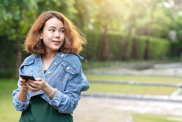 Foto sincero de jóvenes asiáticos atractivos y felices con moda morena pelo rizado elegante diseñador femenino o influyente de pie en el jardín en el exterior de su casa utilizando el teléfono inteligente mirando a un lado para la publicidad.