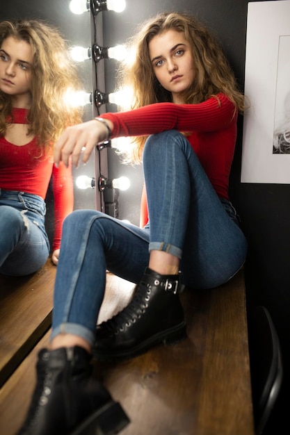 Foto sinnliche junge frau mit lockigem braunem haar, die auf einem holztisch sitzt und in der nähe eines spiegels posiert posing in studio