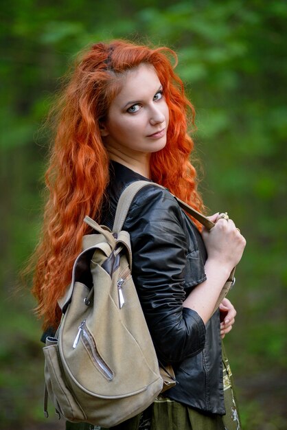 Sinnliche rote Haarfrau der hübschen jungen sexy Mode, die in einem Waldhintergrund aufwirft, kleidete in der Hippie-Rockart-Ausstattung, in der Lederjacke und im Touristenrucksack an.