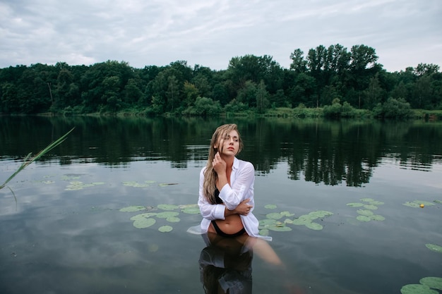 Sinnliches blondes Mädchen mit nassen Haaren in einem weißen Hemd und schwarzem Badeanzug, das nach Regen im See posiert.