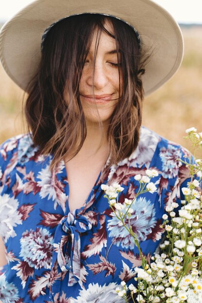 Foto sinnliches porträt eines schönen mädchens mit großem gänseblümchen-bouquet im windigen feld sommer auf dem land schöne junge frau in blauem vintage-kleid und hut mit weißen wildblumen auf der wiese